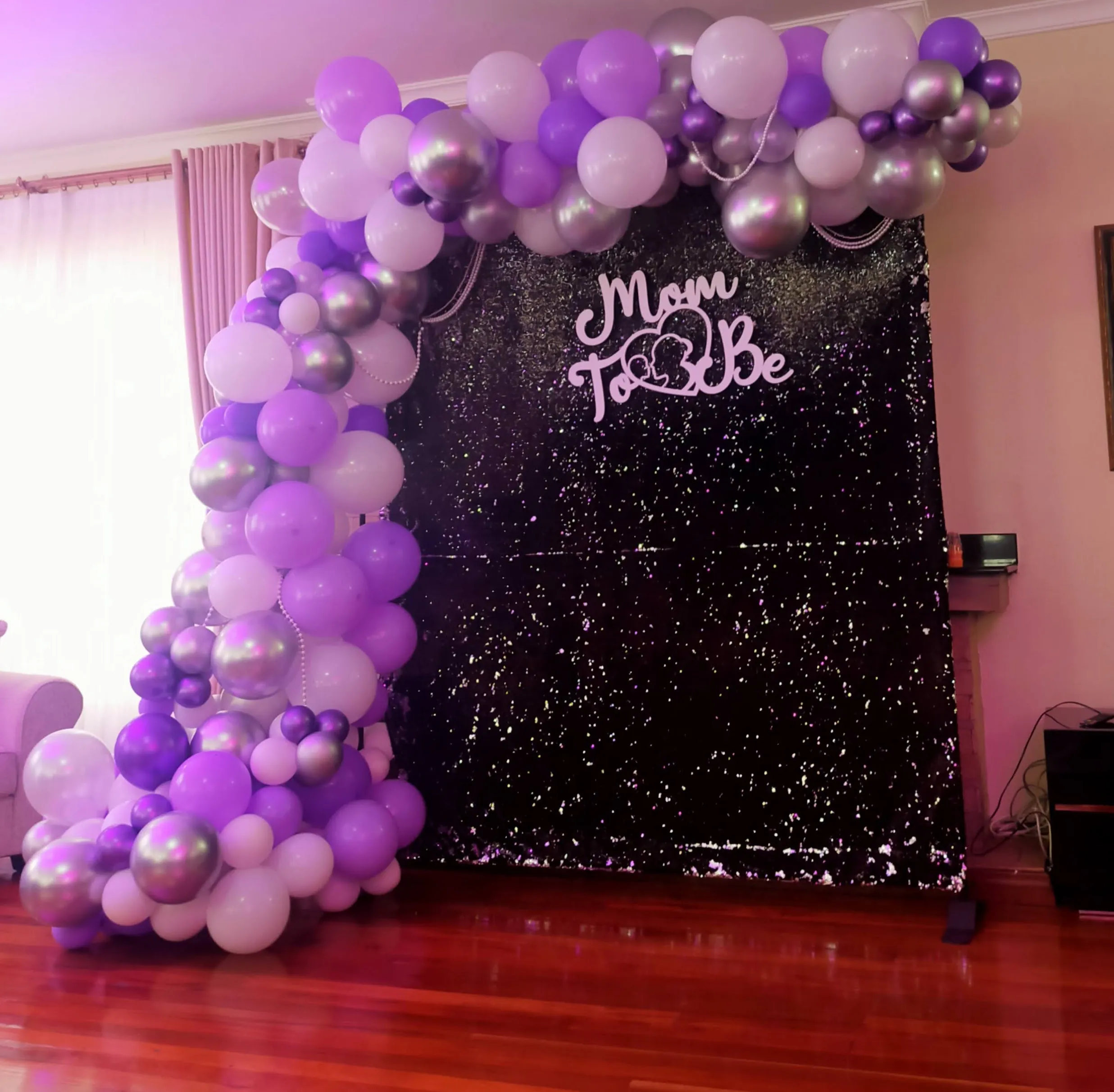 Purple and silver balloon arch around a black sequin backdrop reading Mom To Be
