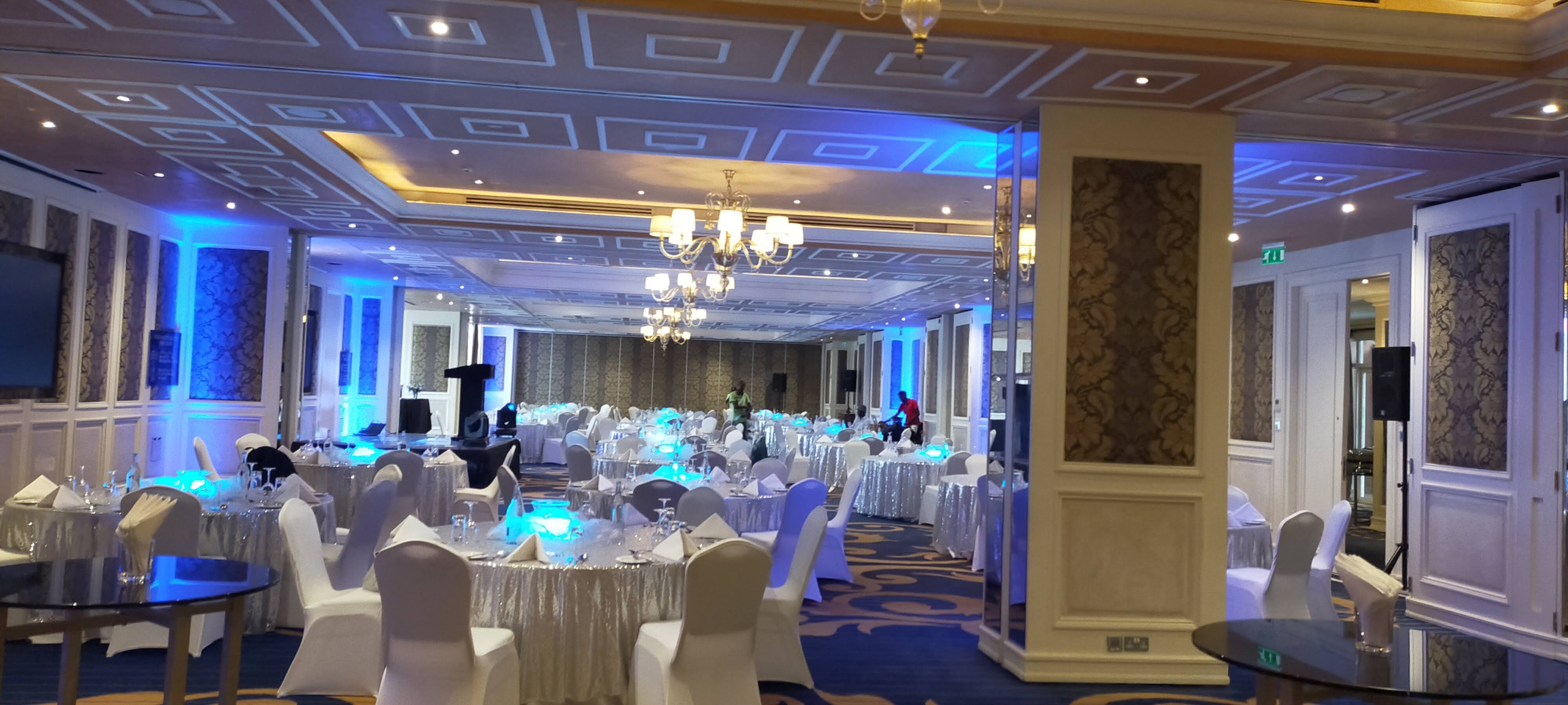 Hotel ballroom with silver-sequinned round tables, white chairs and blue uplighting