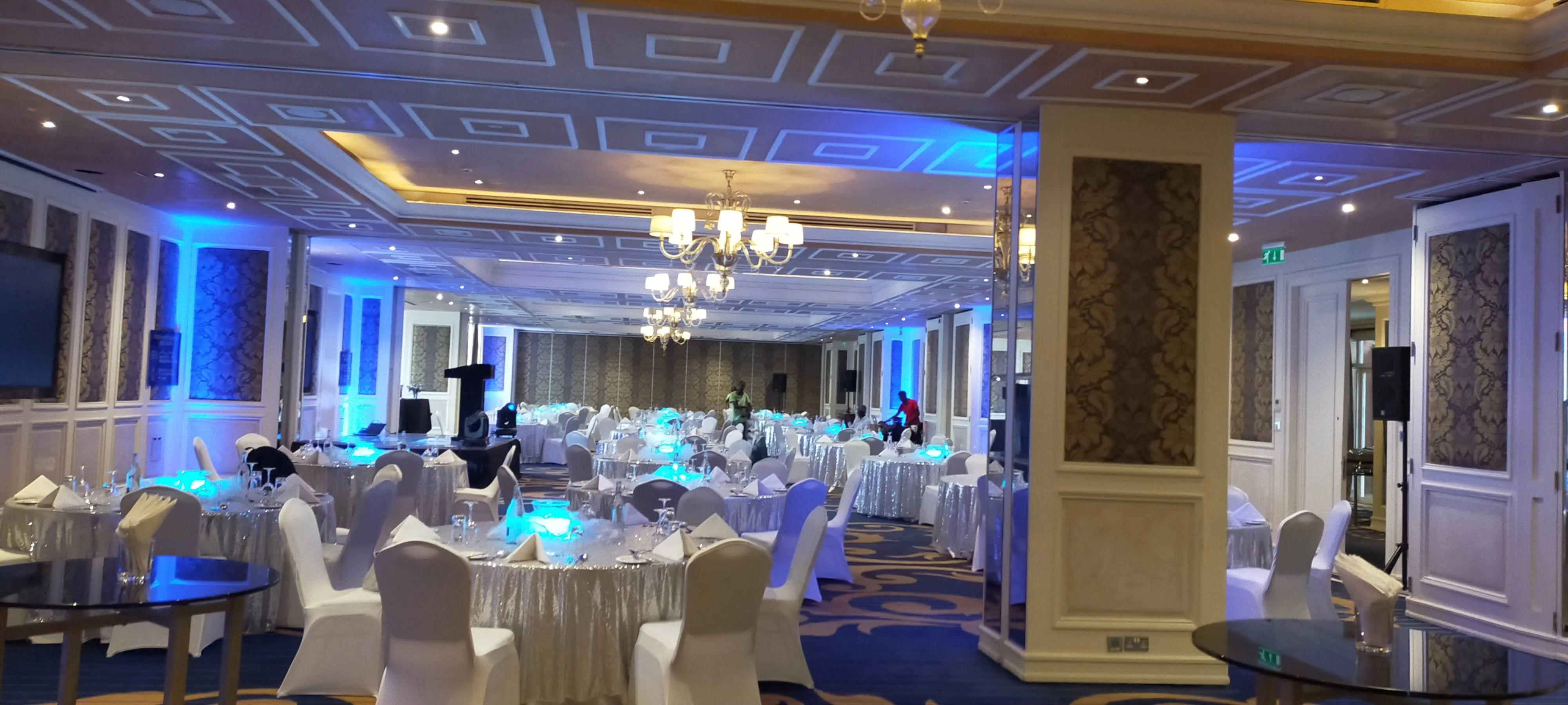 Hotel ballroom with silver-sequinned round tables, white chairs and blue uplighting