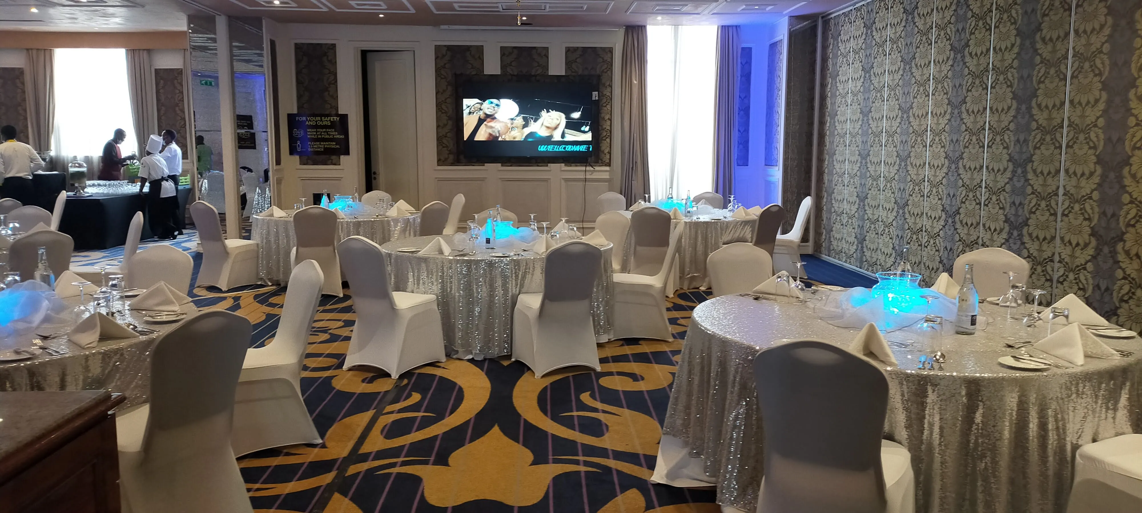 Silver-sequinned banquet tables with white chairs and a welcome screen in a panelled ballroom