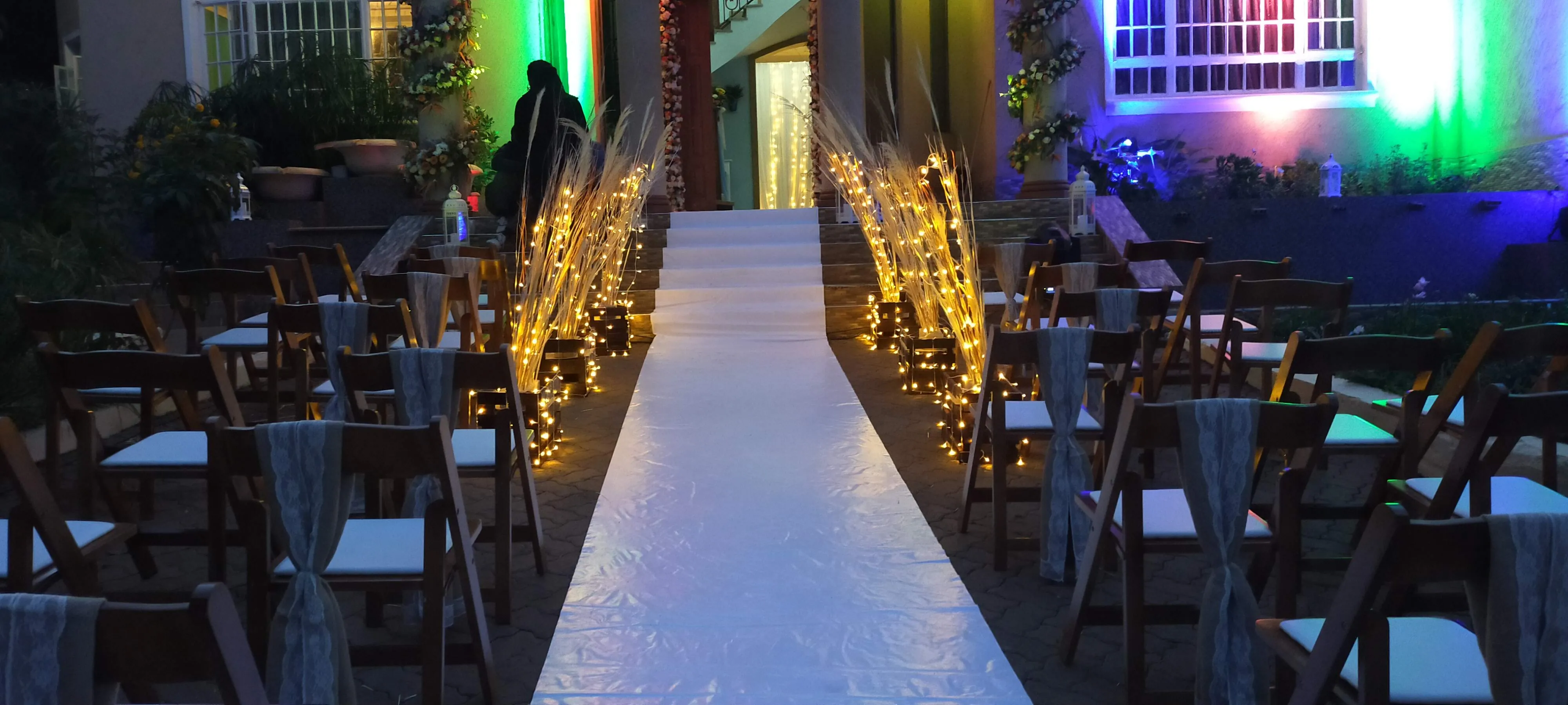 White aisle runner at dusk lined with fairy-light bundles and folding chairs