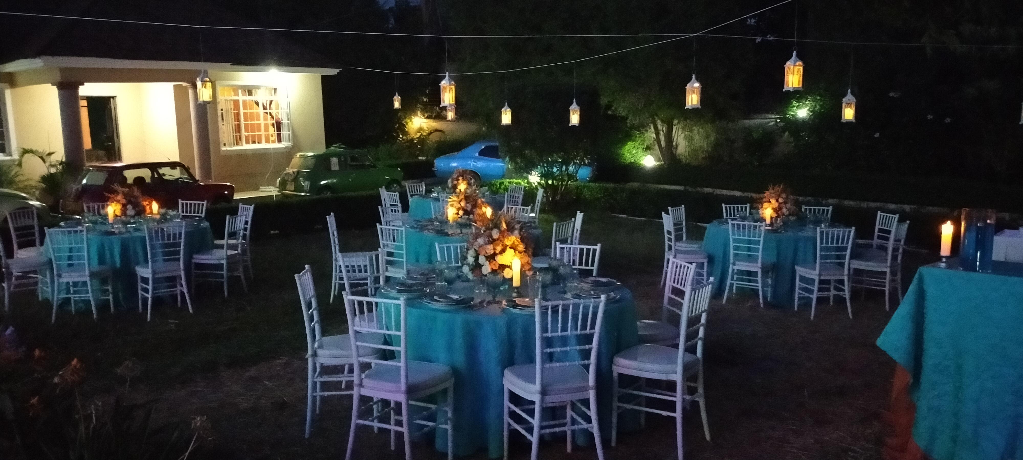 A lantern-lit garden reception at night, teal-clothed round tables glowing under hanging lights